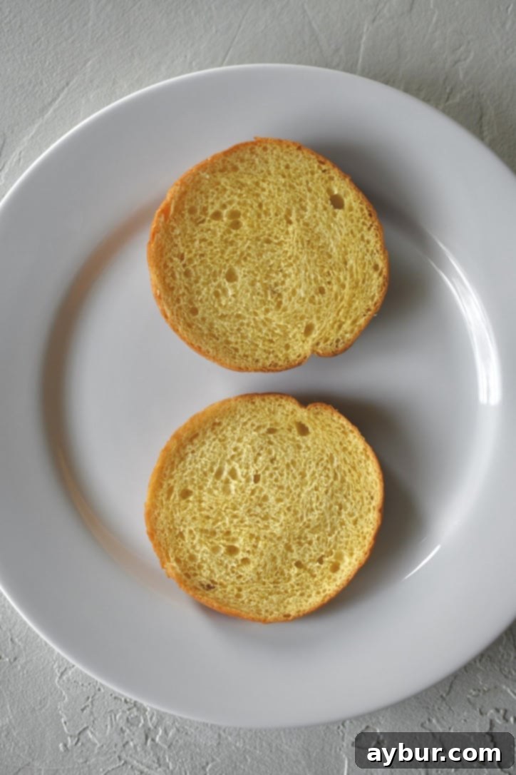 The initial step of assembling a roast beef sandwich, showing a golden, toasted brioche bun placed on a plate, ready for the toppings.