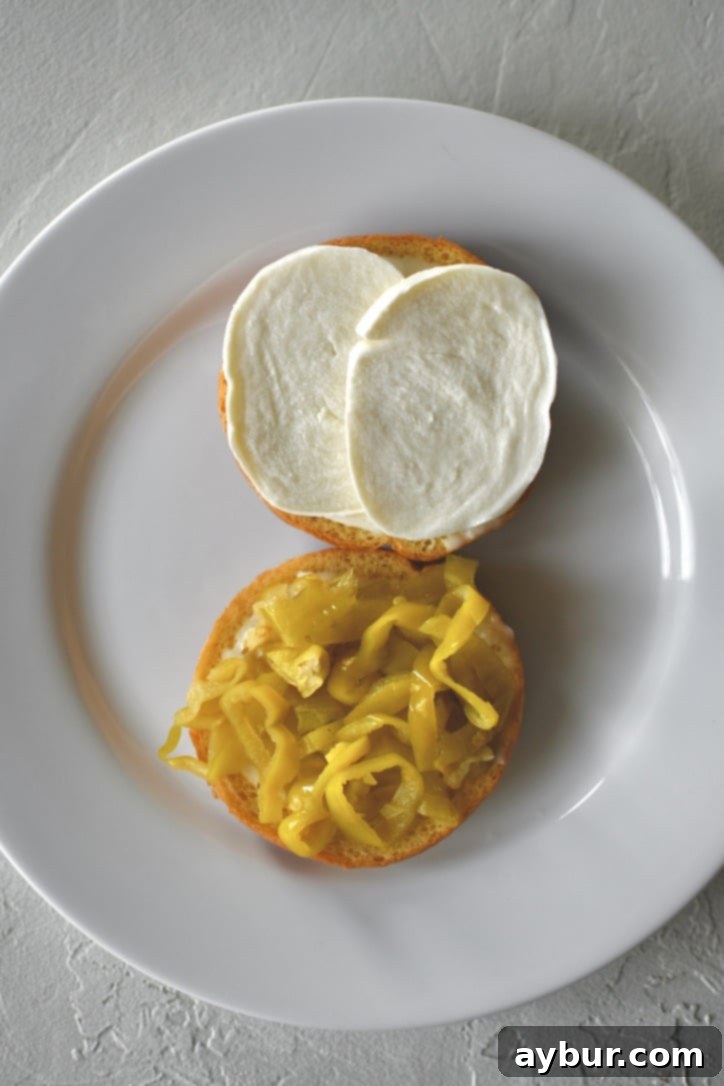 The layering process of a roast beef sandwich, showing banana peppers and creamy mozzarella cheese being carefully placed onto the toasted bun, already spread with horseradish sauce.