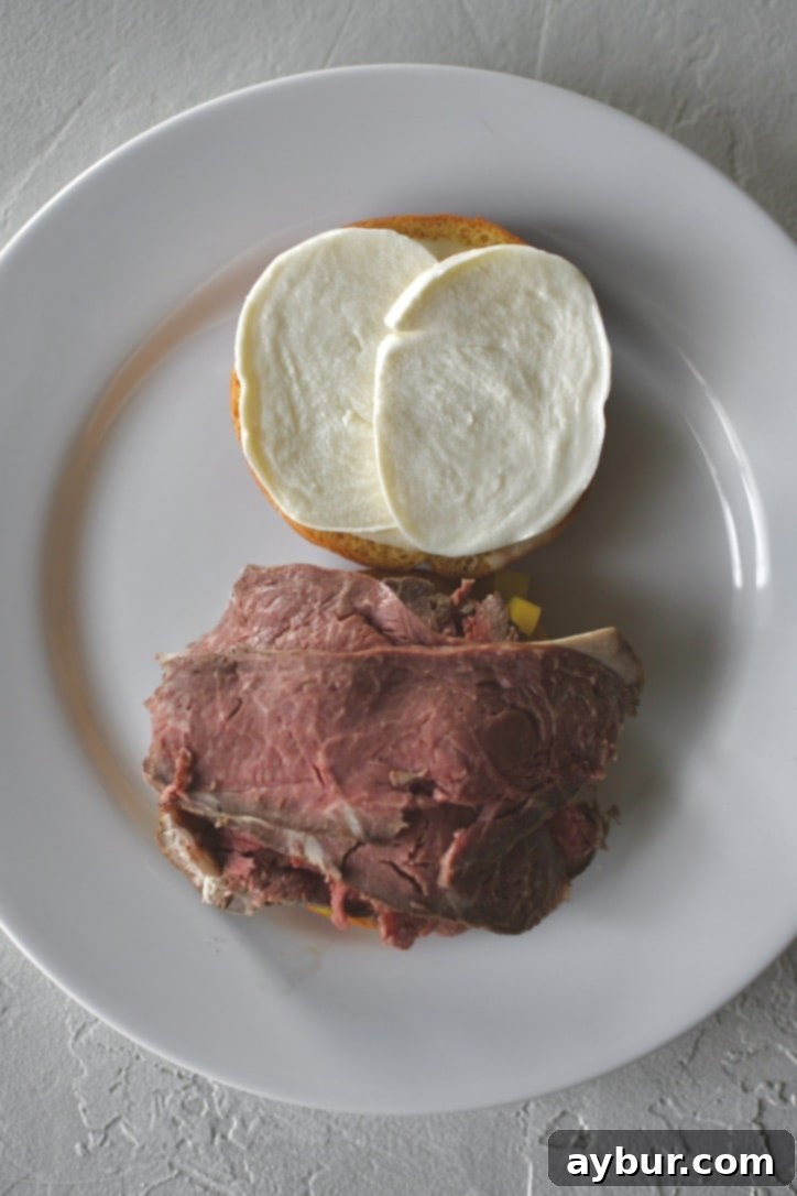 The next stage of sandwich assembly, featuring tender slices of carved roast beef being added generously on top of the banana peppers and horseradish-coated bun.