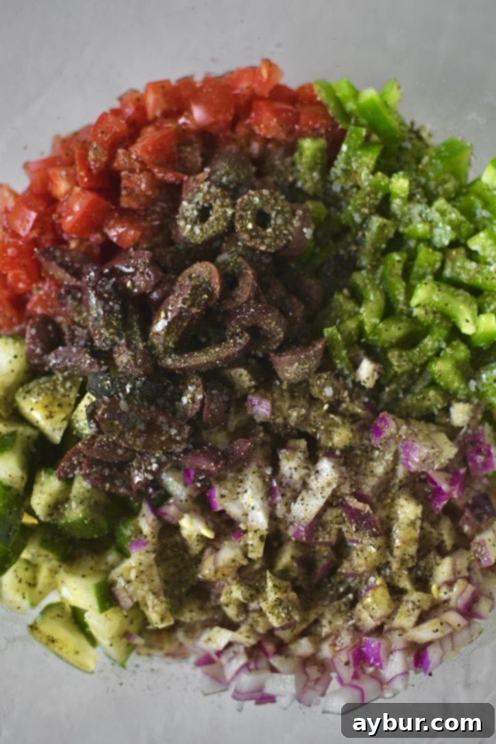 A hand drizzling red wine vinegar and olive oil over the chopped vegetables in a bowl, alongside Greek seasoning, salt, and pepper.