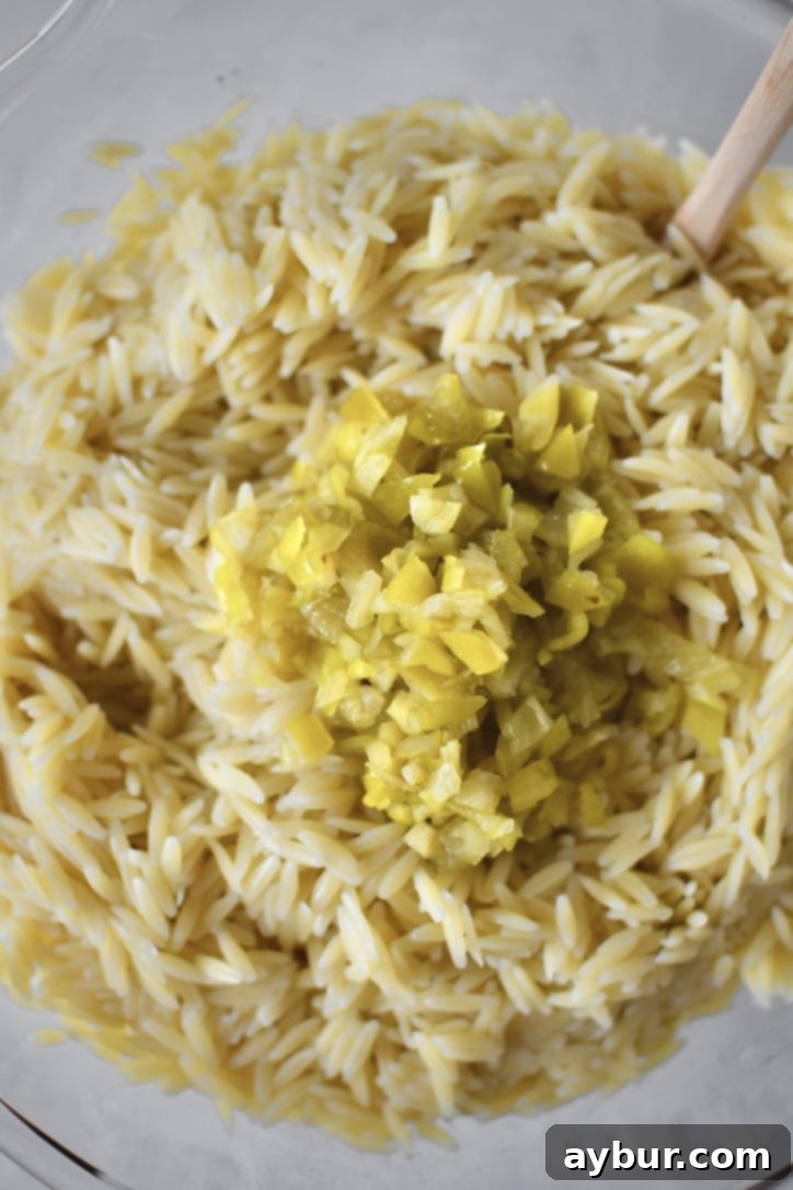 Cooked and cooled orzo pasta being added to a bowl of seasoned vegetables, along with pepperoncini peppers, for the Greek Pasta Salad.