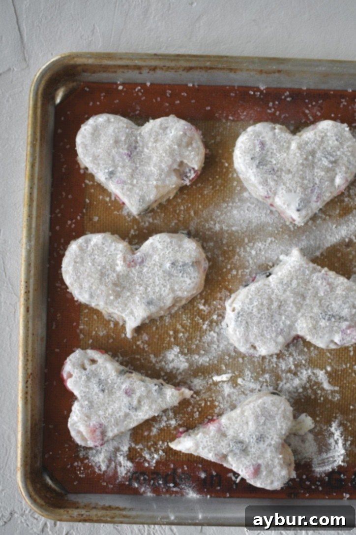Chilled heart-shaped scones brushed with heavy cream and generously topped with sanding sugar on a baking sheet.