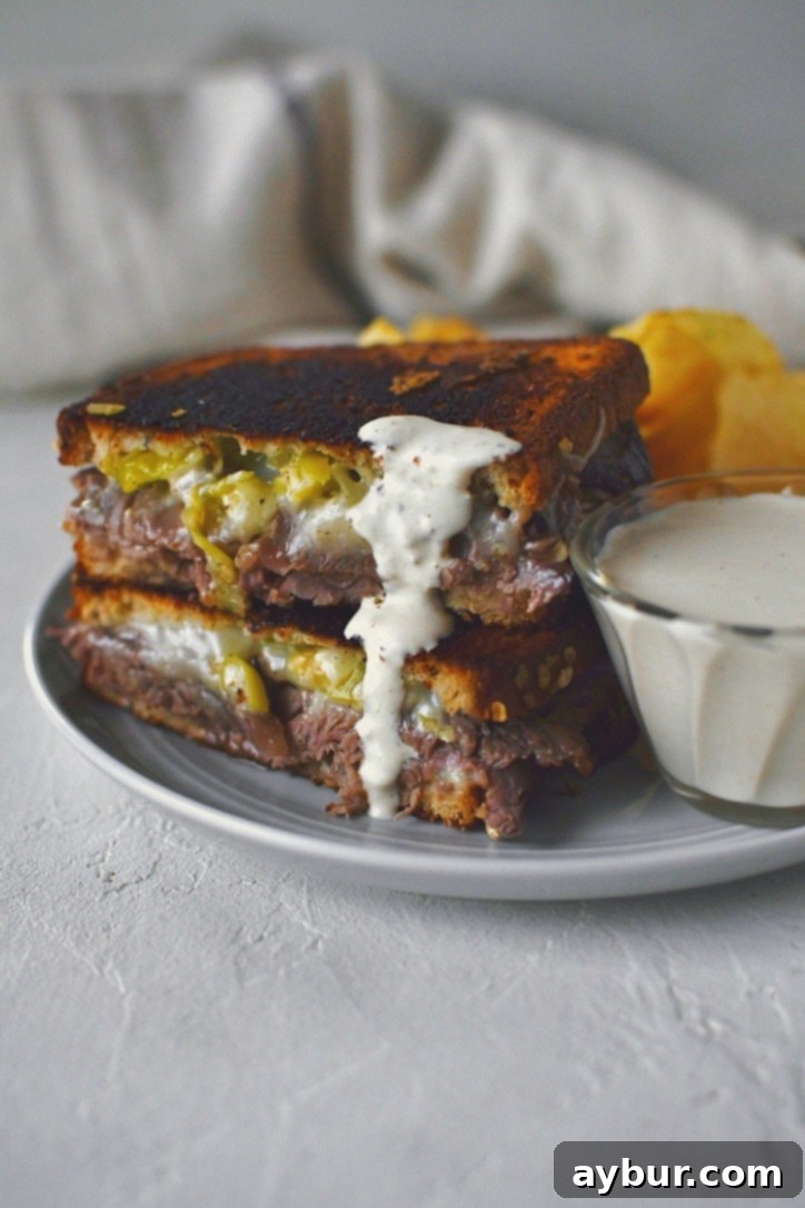 A savory roast beef grilled cheese sandwich, perfectly toasted, with a corner dipped into a small bowl of creamy horseradish sauce. The rich texture of the sauce is visible as it clings to the sandwich.
