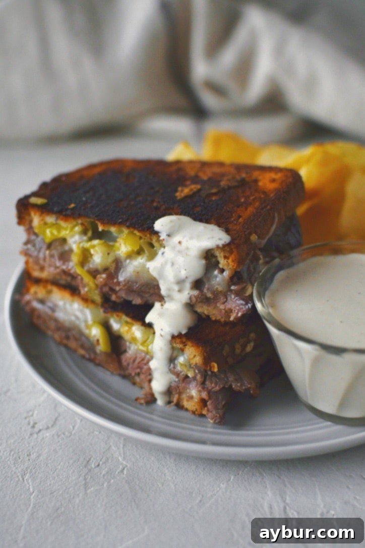 A golden-brown roast beef grilled cheese sandwich, cut in half diagonally, with a generous portion of horseradish cream sauce waiting for a dip. The inviting steam rises from the warm sandwich.