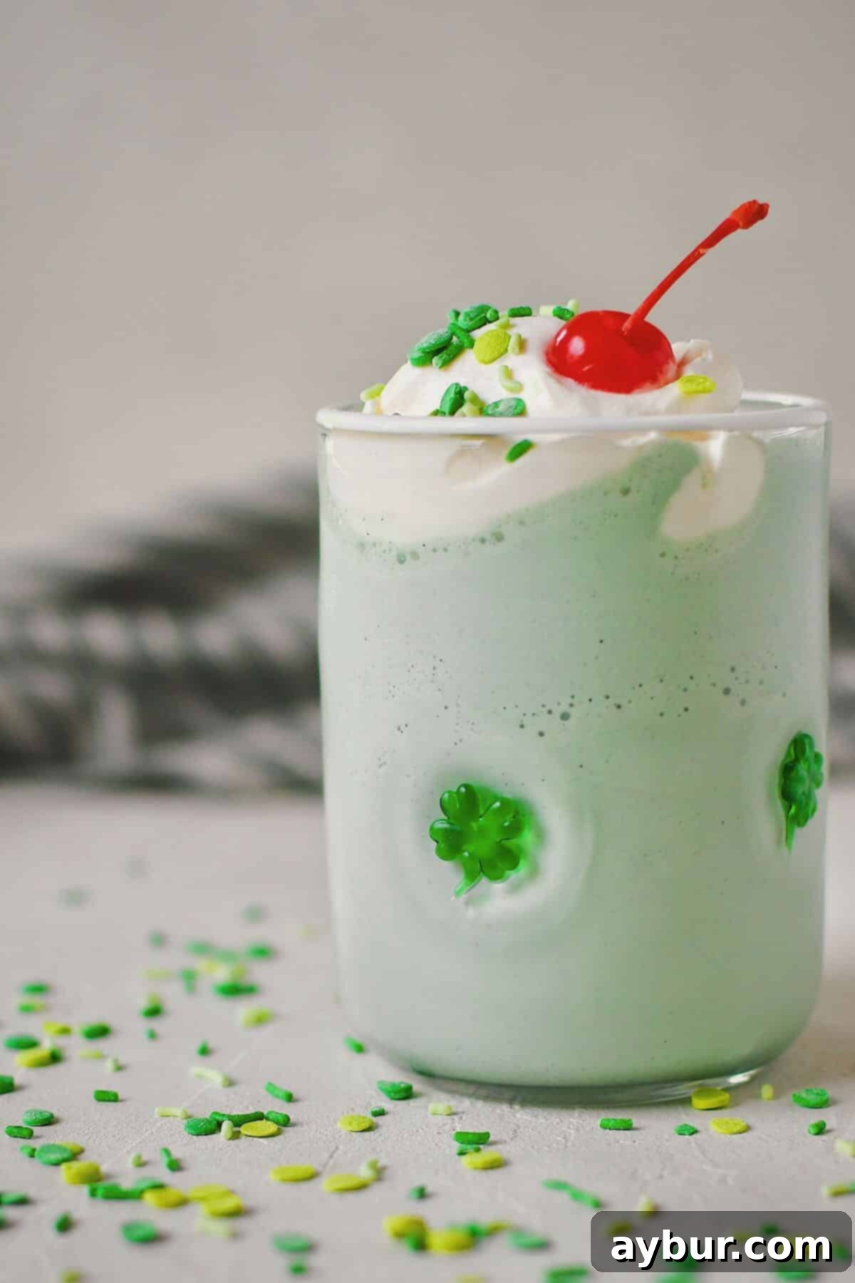Vibrant green Shamrock Shake in a festive shamrock-patterned glass, generously topped with fluffy homemade whipped cream, colorful sprinkles, and a bright red cherry.