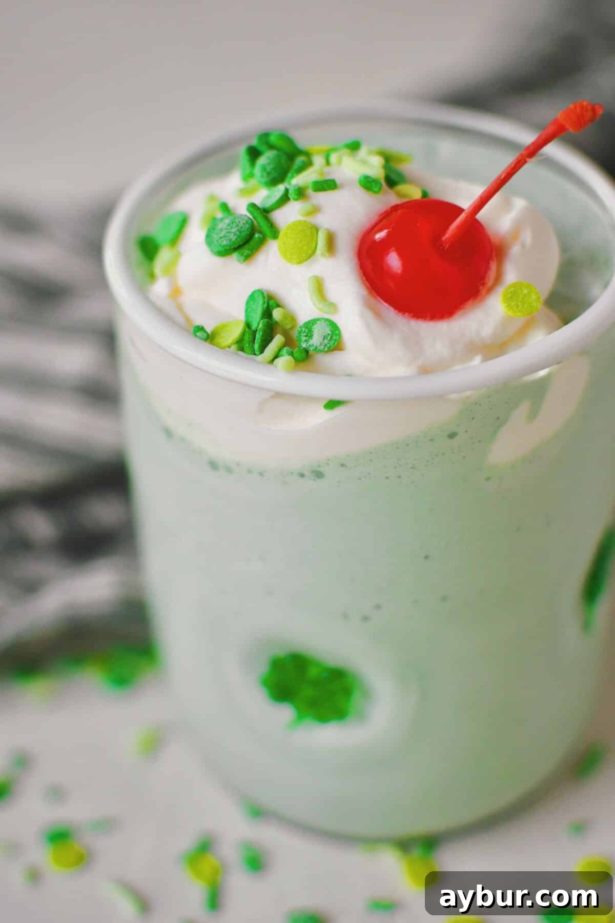 Close-up of a delectable Shamrock Shake in a shamrock-themed glass, artfully crowned with a generous swirl of whipped cream, vibrant green sprinkles, and a single red cherry. The shake's smooth, green texture is clearly visible.