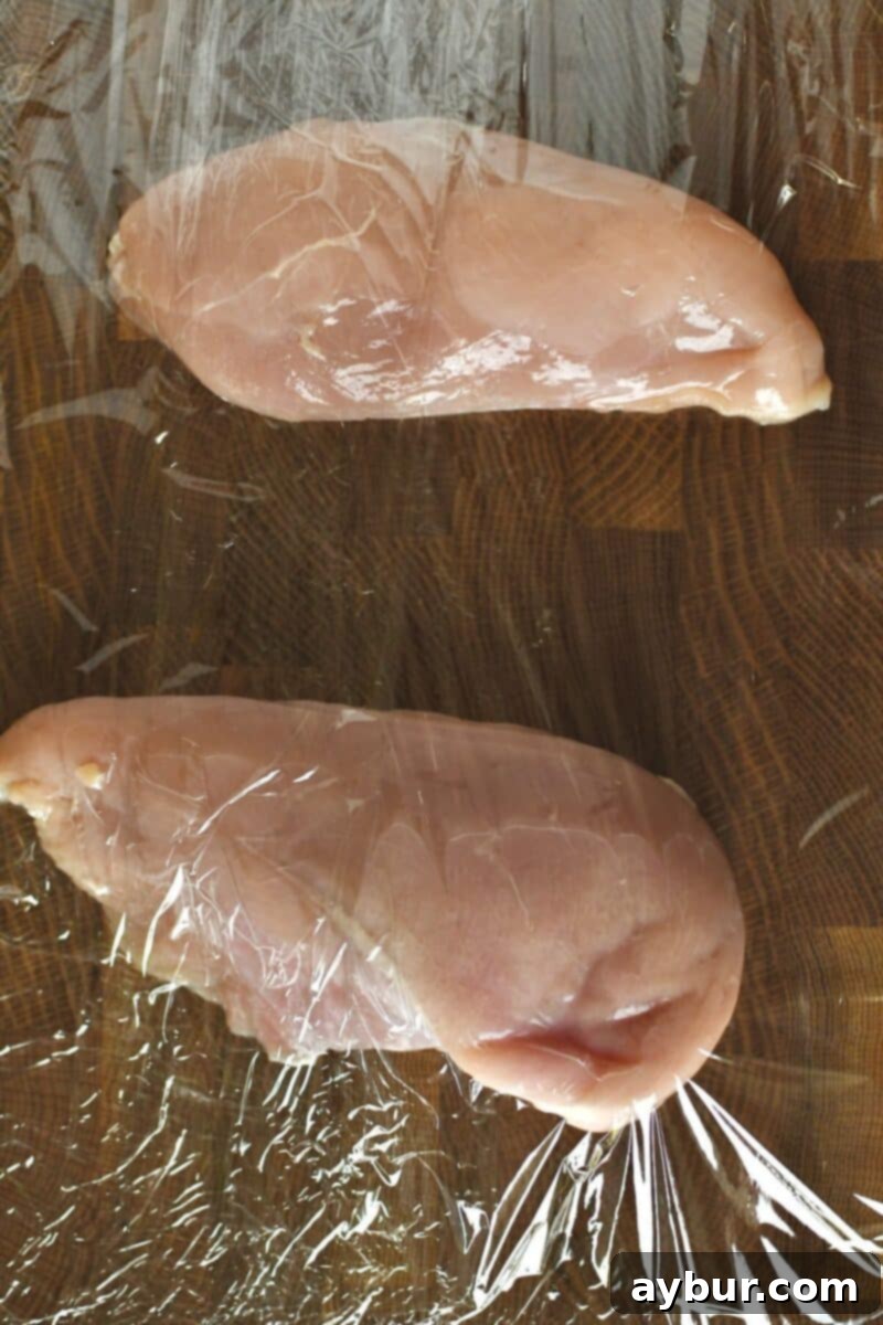Raw chicken breasts on a cutting board, covered with cling film, before being tenderized to an even thickness.