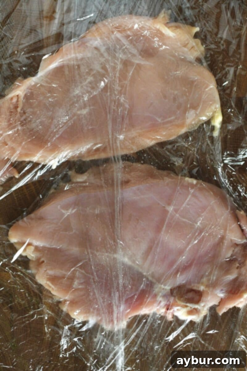 Chicken breasts on a cutting board, evenly flattened under cling film after tenderizing with a meat mallet.