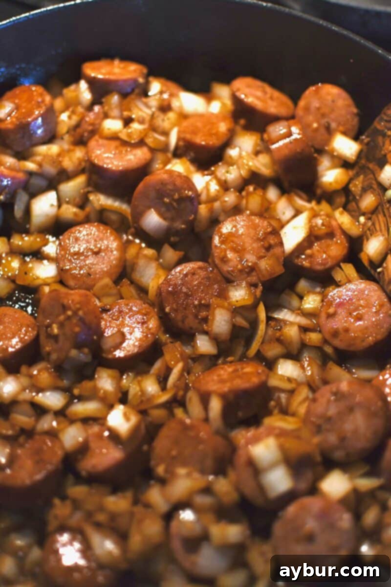 Diced onions, minced garlic, and sliced Andouille sausage cooking together in a skillet, browning nicely.