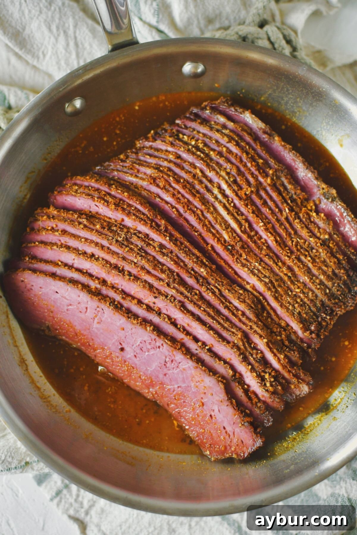 Sliced homemade pastrami being gently reheated in a skillet with its reserved cooking juices, under a lid.