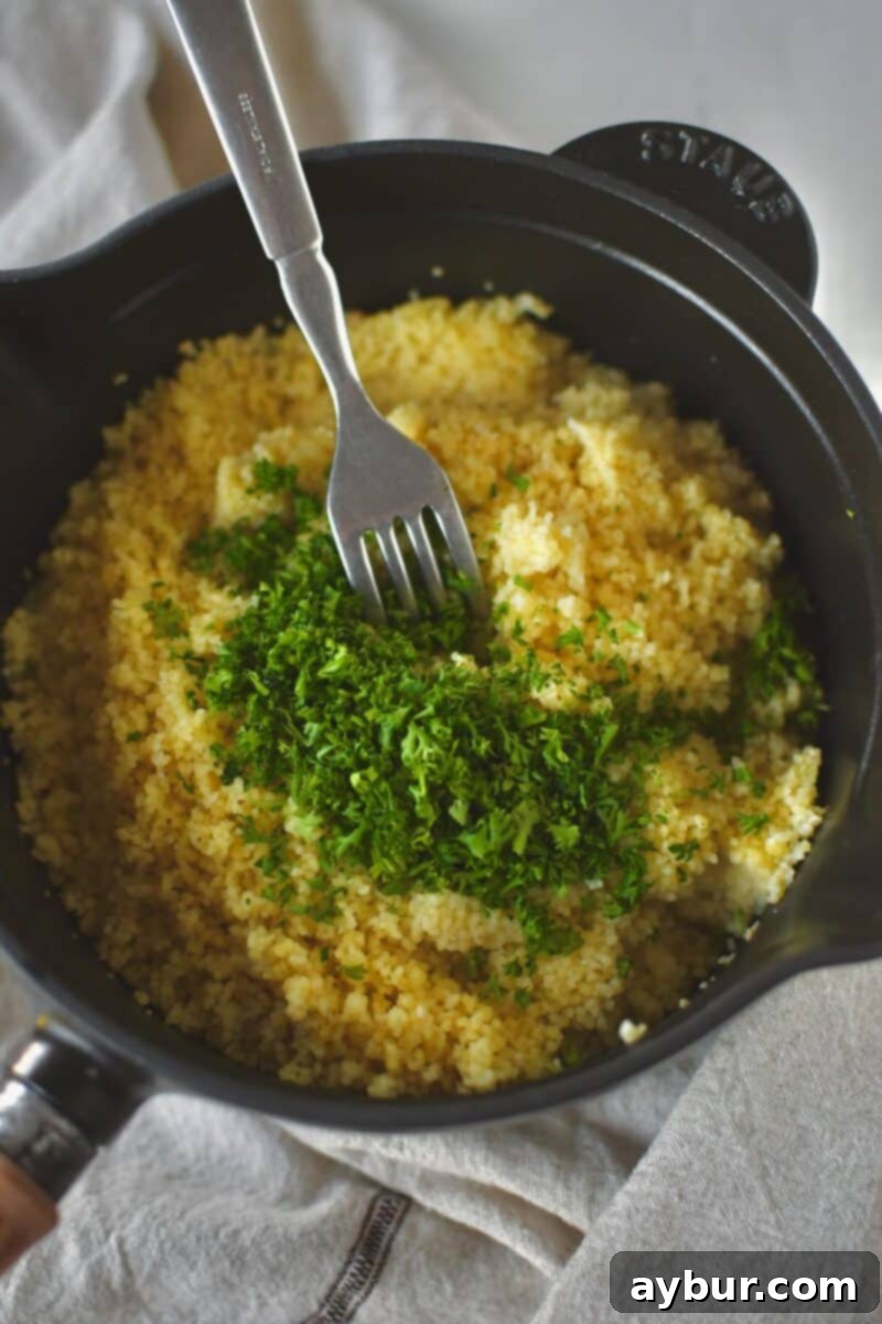 Cooked couscous being fluffed with a fork, with chopped fresh parsley added and gently folded in, creating a light and airy texture.