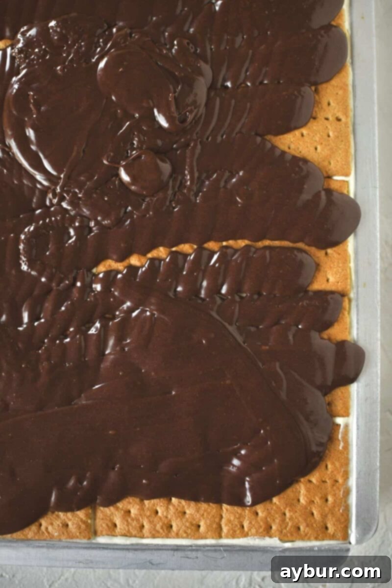 The moment of truth: warm, glossy chocolate frosting being poured over the final layer of graham crackers, preparing to spread.