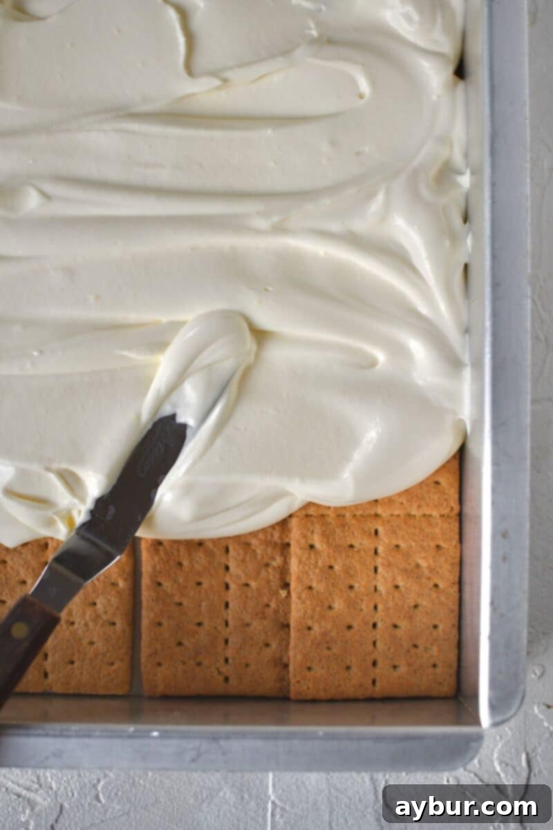 A generous amount of the creamy pudding mixture being spread smoothly over the first layer of graham crackers in the baking pan.