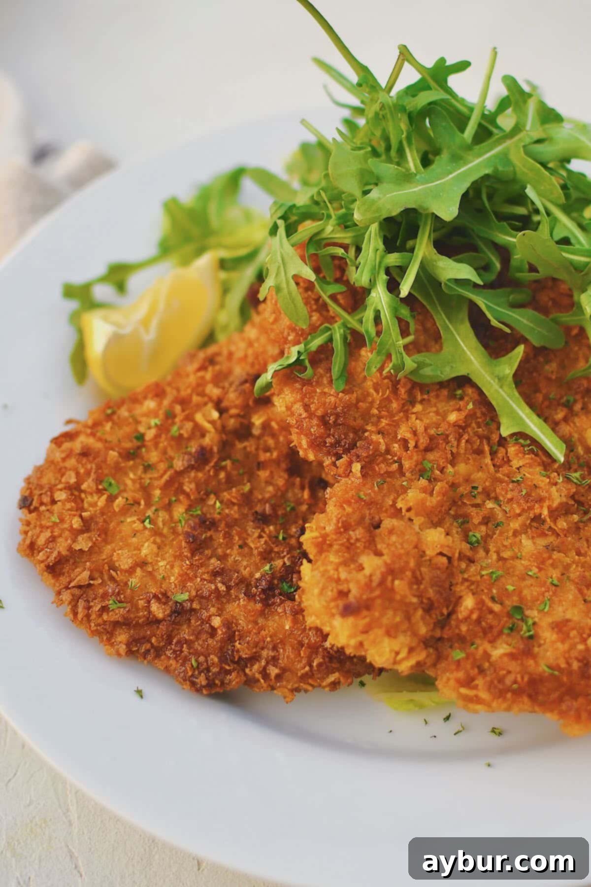 Pan-Fried Pork Perfection 2 Crispy Pork Schnitzel served on a white plate with a fresh arugula salad and bright lemon wedges, ready to be enjoyed.