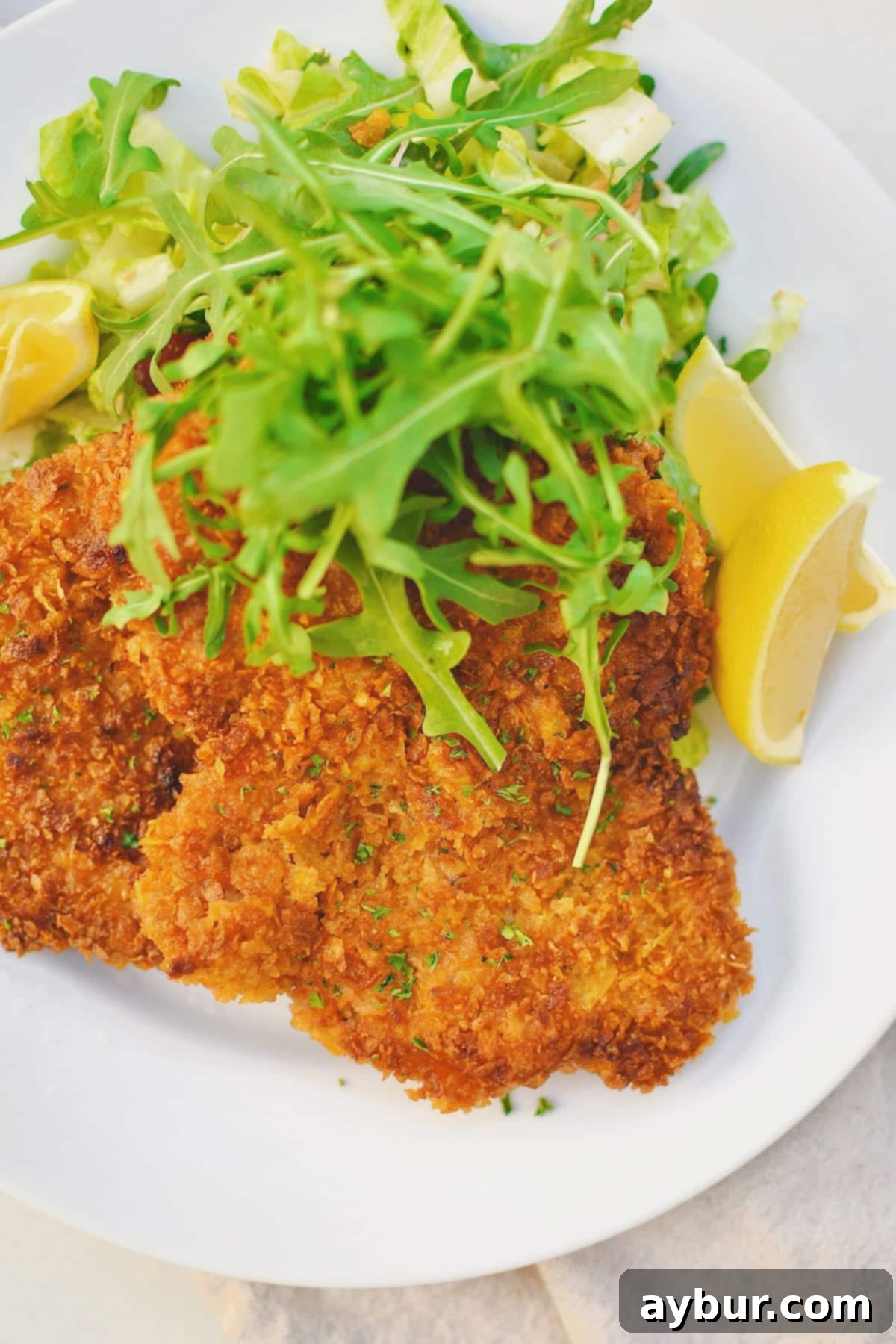 Pan-Fried Pork Perfection 9 A beautifully golden-brown Pork Schnitzel on a white plate, served with a fresh arugula salad and lemon wedges, ready to be enjoyed.