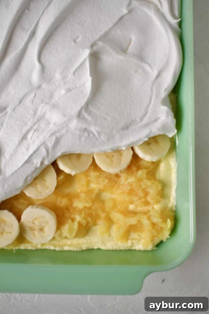 Ultimate Banana Split Cake 9 The final stages of assembling the Banana Split Cake: spreading out the crushed pineapple over the creamy filling, adding a fresh layer of sliced bananas, and then topping everything with a smooth layer of whipped cream.