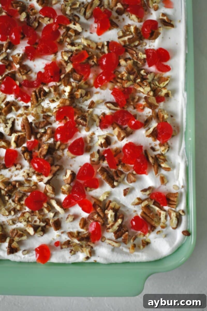 Ultimate Banana Split Cake 10 The fully assembled Banana Split Cake in the baking dish, ready to be chilled. It's garnished with vibrant chopped maraschino cherries and a generous sprinkle of toasted pecans on top.