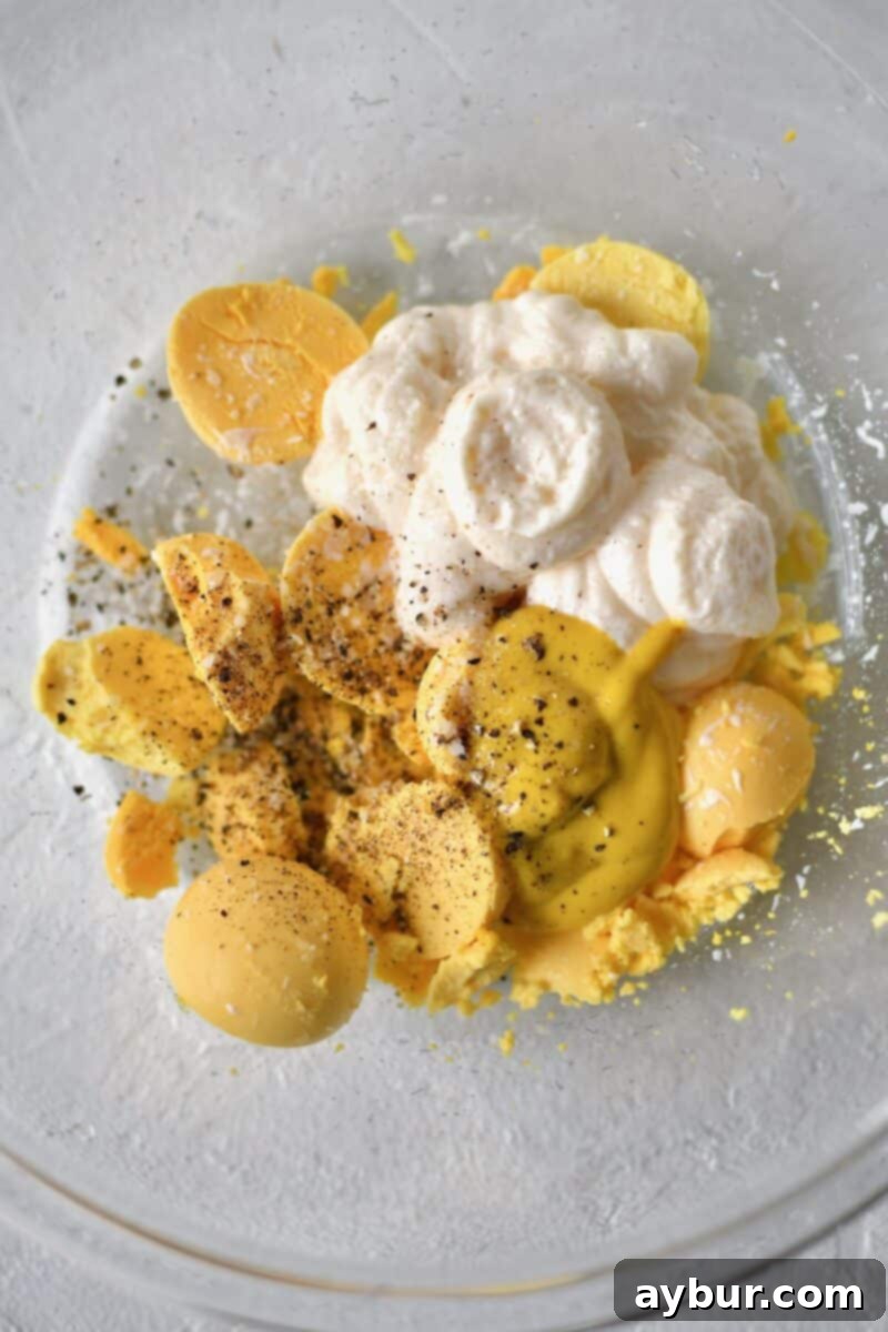 A small bowl containing vibrant yellow egg yolks, now enhanced with dollops of creamy mayonnaise, a squeeze of yellow mustard, a sprinkle of salt, and a dash of freshly ground black pepper, awaiting the mashing process for the deviled egg filling.