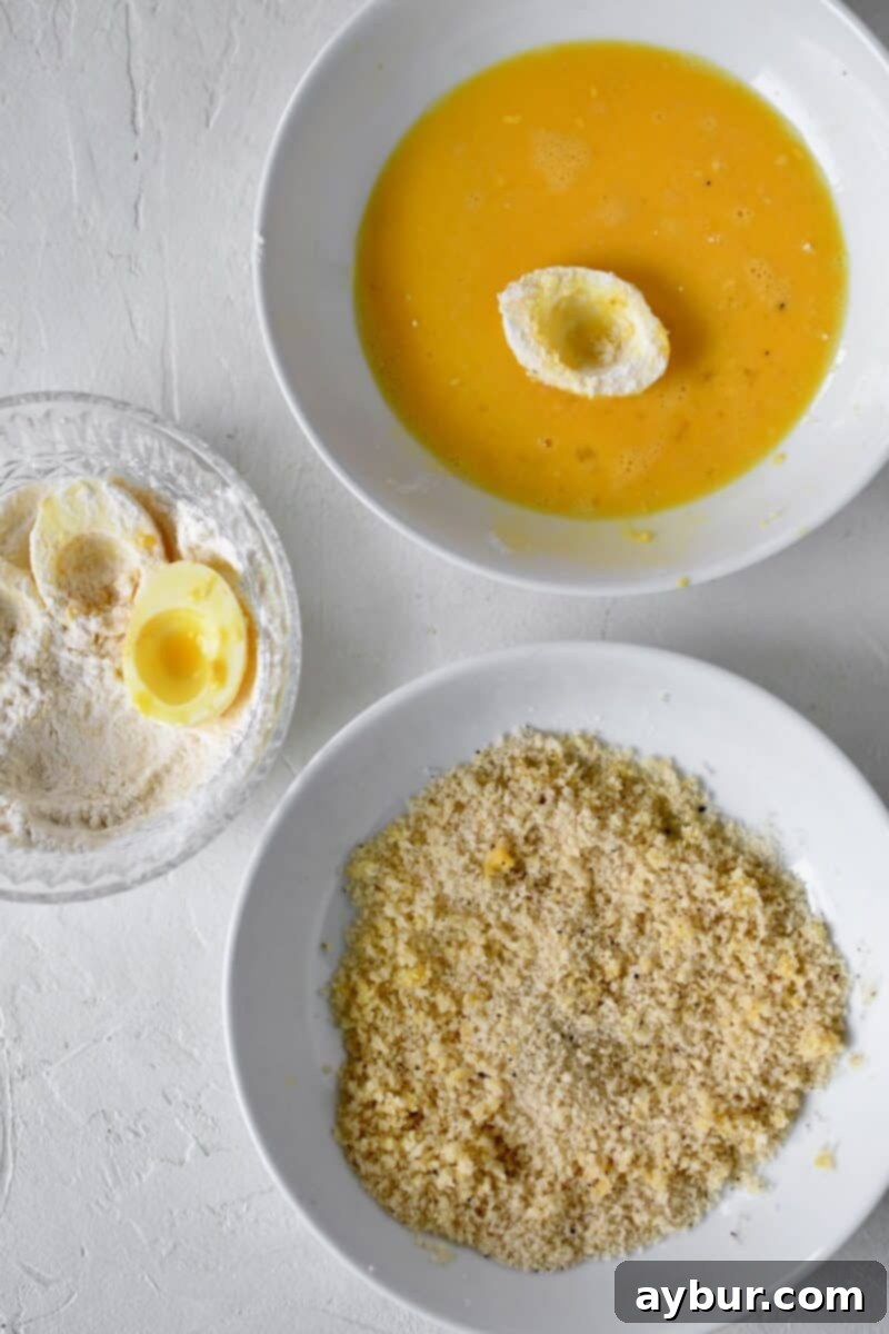 A comprehensive view of the breading station set up for deep-fried deviled eggs, showing the sequence of dusting an egg white in flour, dipping it into a bowl of beaten egg wash, and finally dredging it generously in a bowl of panko bread crumbs, ready for frying.