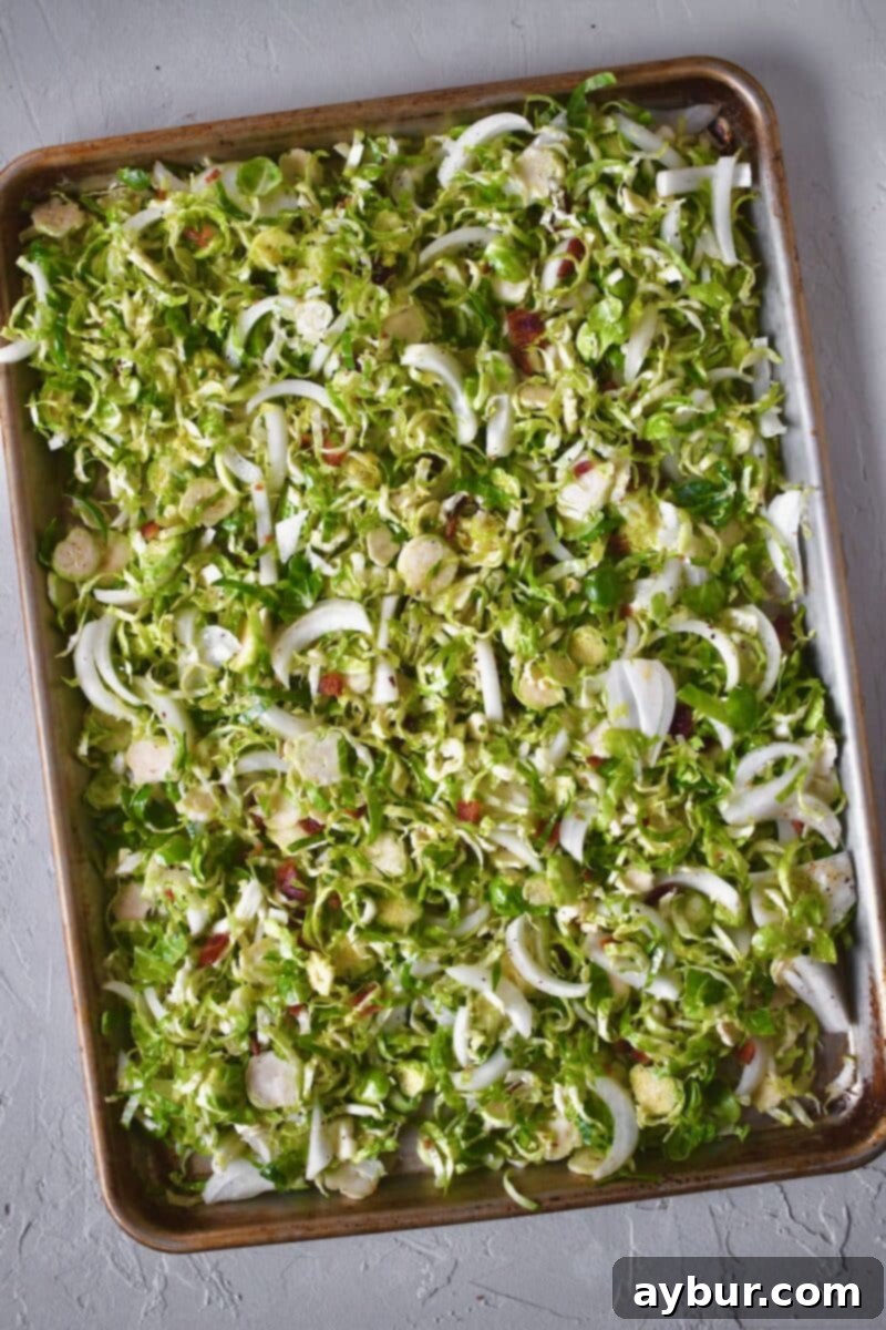 Shaved Brussels Sprouts on a baking sheet seasoned with the oil, bacon fat, salt, and pepper, and tossed with onion slices ready to be cooked.