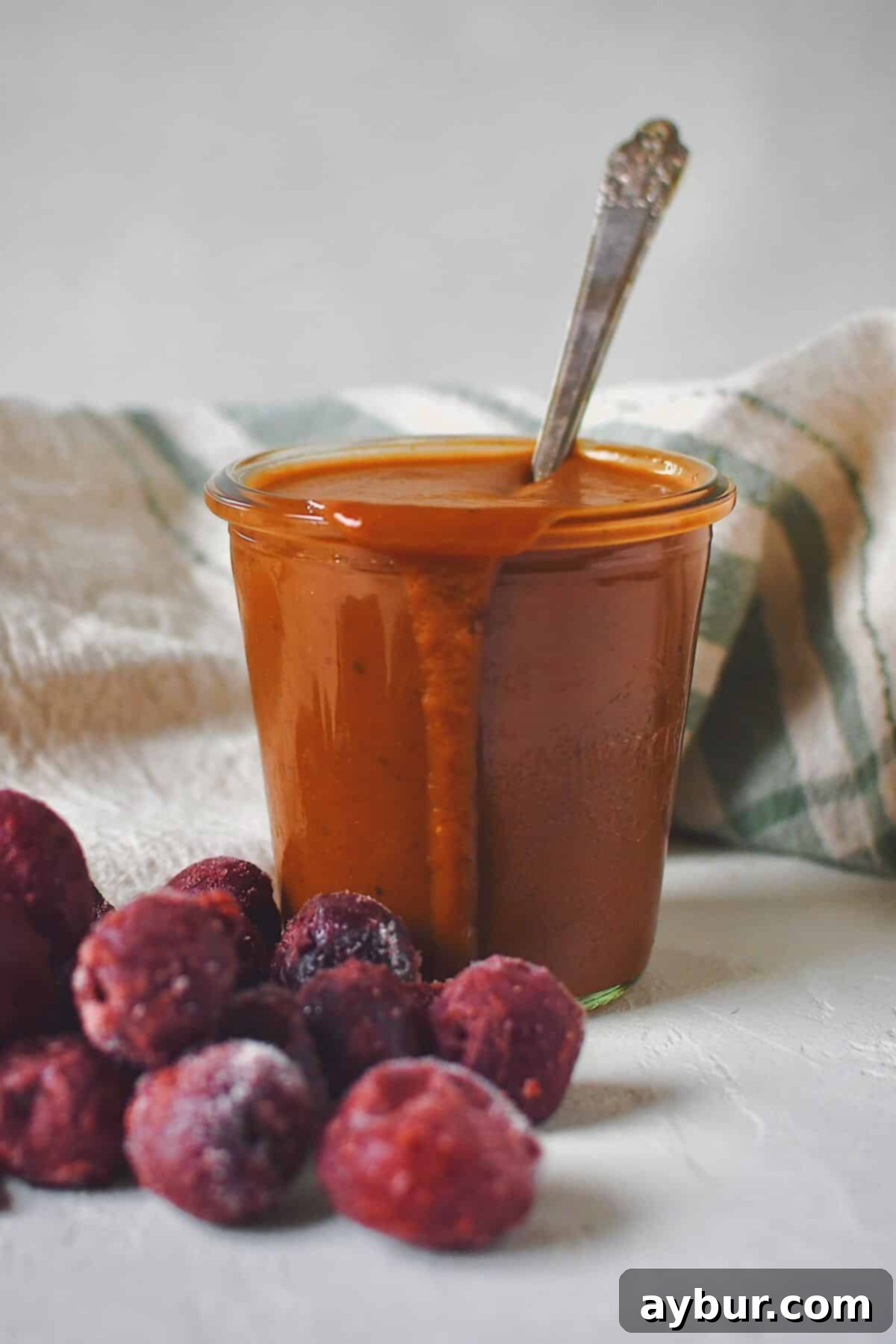 A jar of finished Dr. Pepper BBQ Sauce with a spoon in it, surrounded by frozen tart cherries, ready for serving.