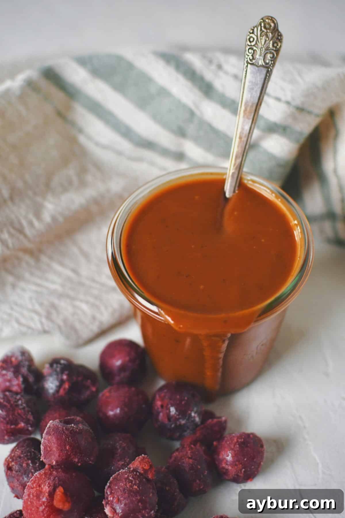 Another beautiful shot of the finished Dr. Pepper BBQ Sauce in a glass jar, with a spoon nestled inside and fresh frozen cherries scattered in front.