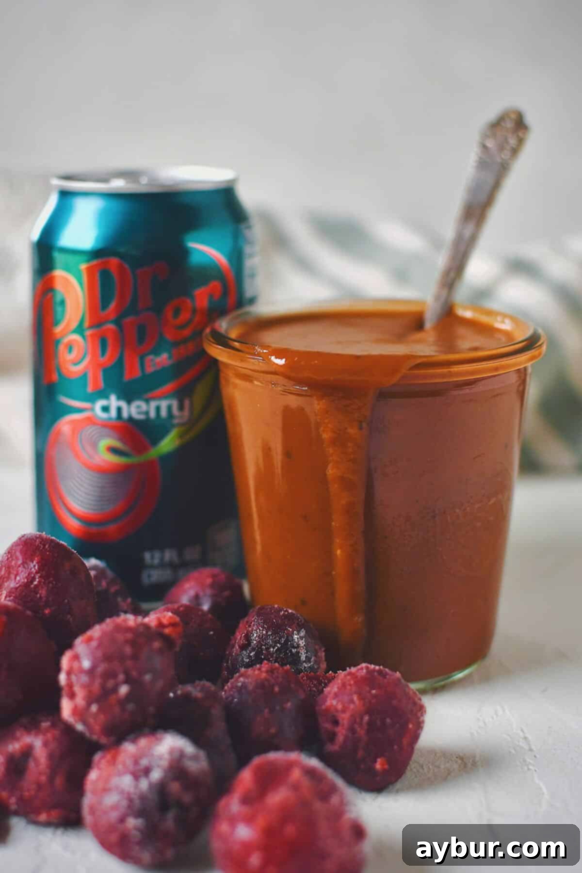 A jar of finished Dr. Pepper BBQ Sauce with a spoon in it, a can of cherry Dr. Pepper in the background, and frozen cherries in the foreground, showcasing the key ingredients.