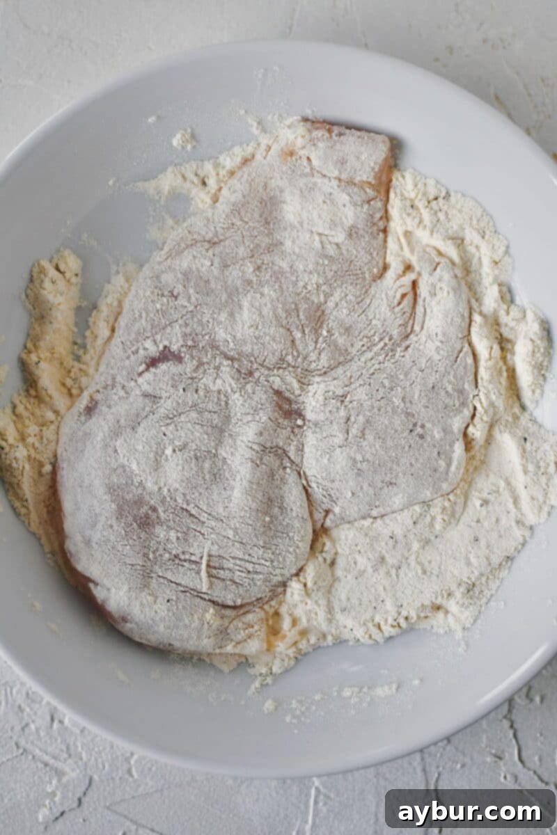 Velvety Mushroom Chicken 4 Tenderized chicken breast being dredged through a bowl of seasoned flour, preparing for pan-frying.