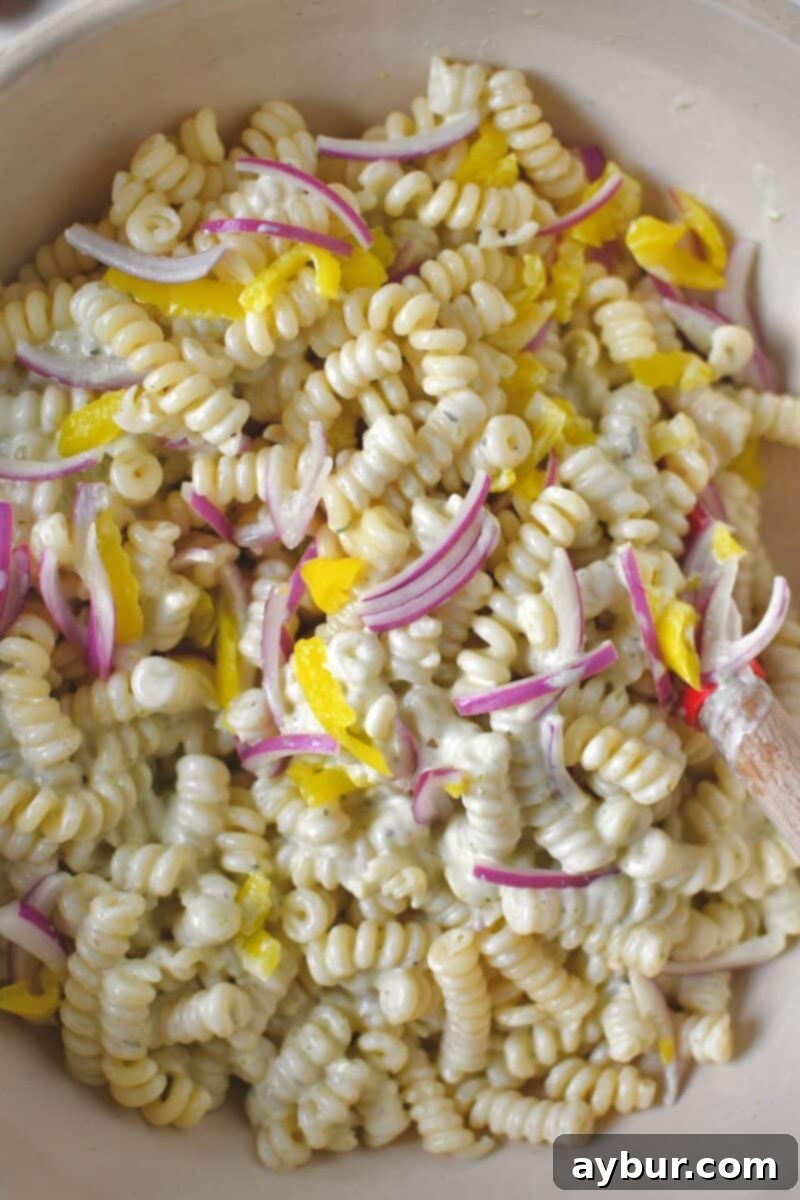 Tossing the cooled pasta, marinated onions, and banana peppers with the creamy pesto ranch dressing in a large bowl.