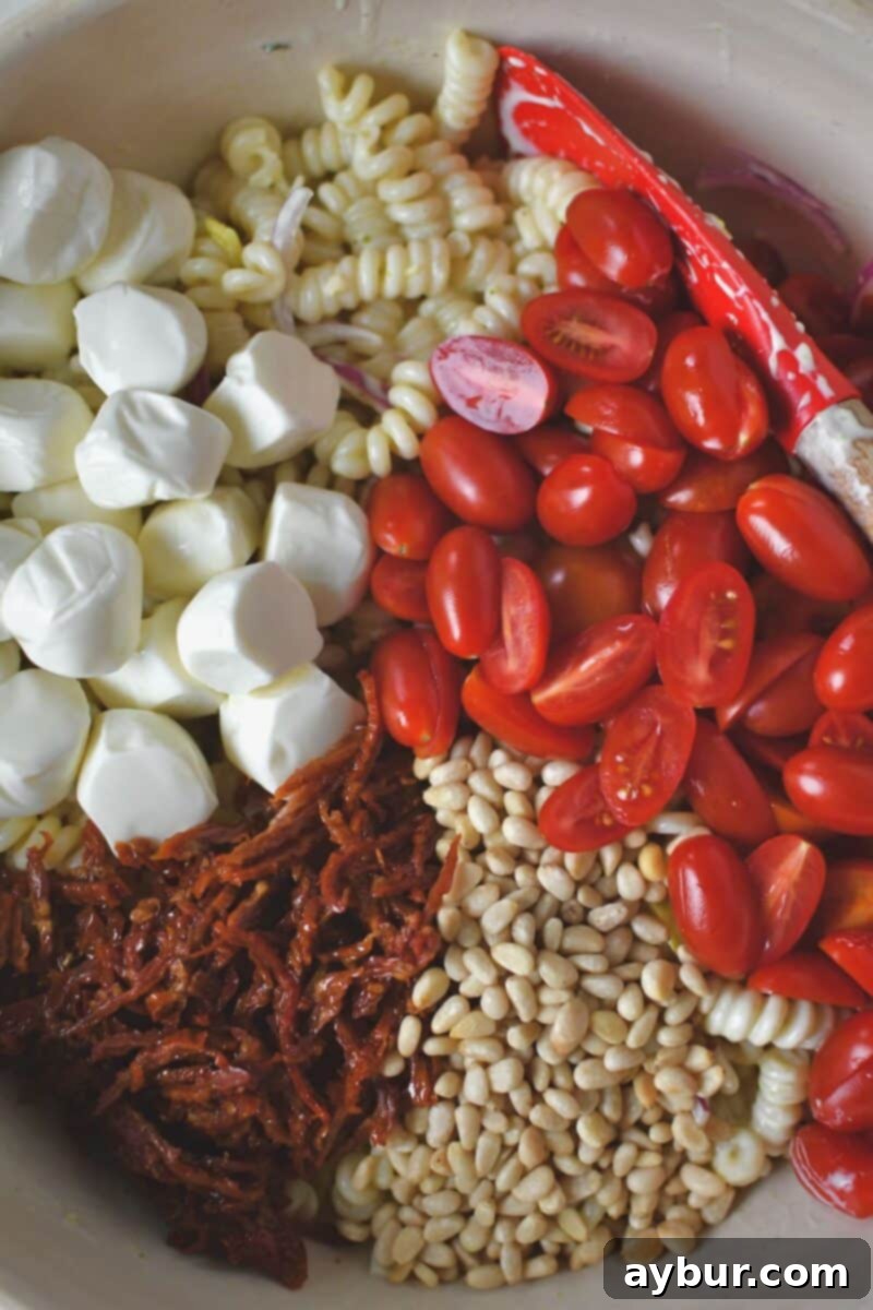 Adding mozzarella balls, halved grape tomatoes, finely sliced sun-dried tomatoes, and toasted pine nuts to the dressed pasta salad.