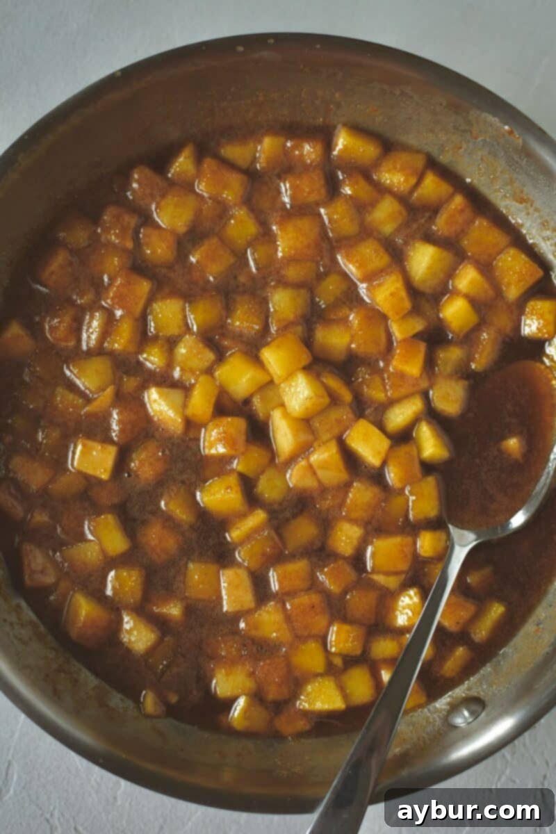 Warm Spiced Apple Pancakes 7 A skillet filled with beautifully cooked diced apples, coated in a glistening, bubbling caramel sauce infused with cinnamon, forming a delectable pancake topping.