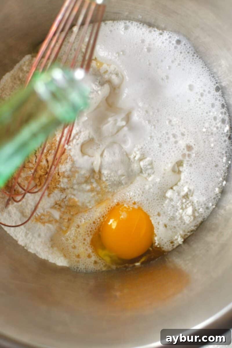 Dry ingredients including flour, baking powder, and roasted garlic powder in a bowl, with an egg, as sparkling water is poured in to form a smooth batter.