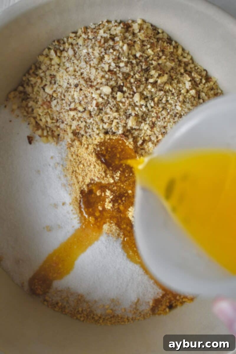 Melted butter being poured into a bowl containing graham cracker crumbs, ground hazelnuts, sugar, and cinnamon for the cheesecake crust.