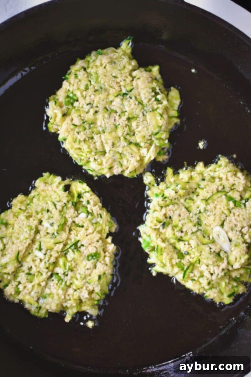 Flattened scoops of zucchini batter frying in a large pan with some oil.