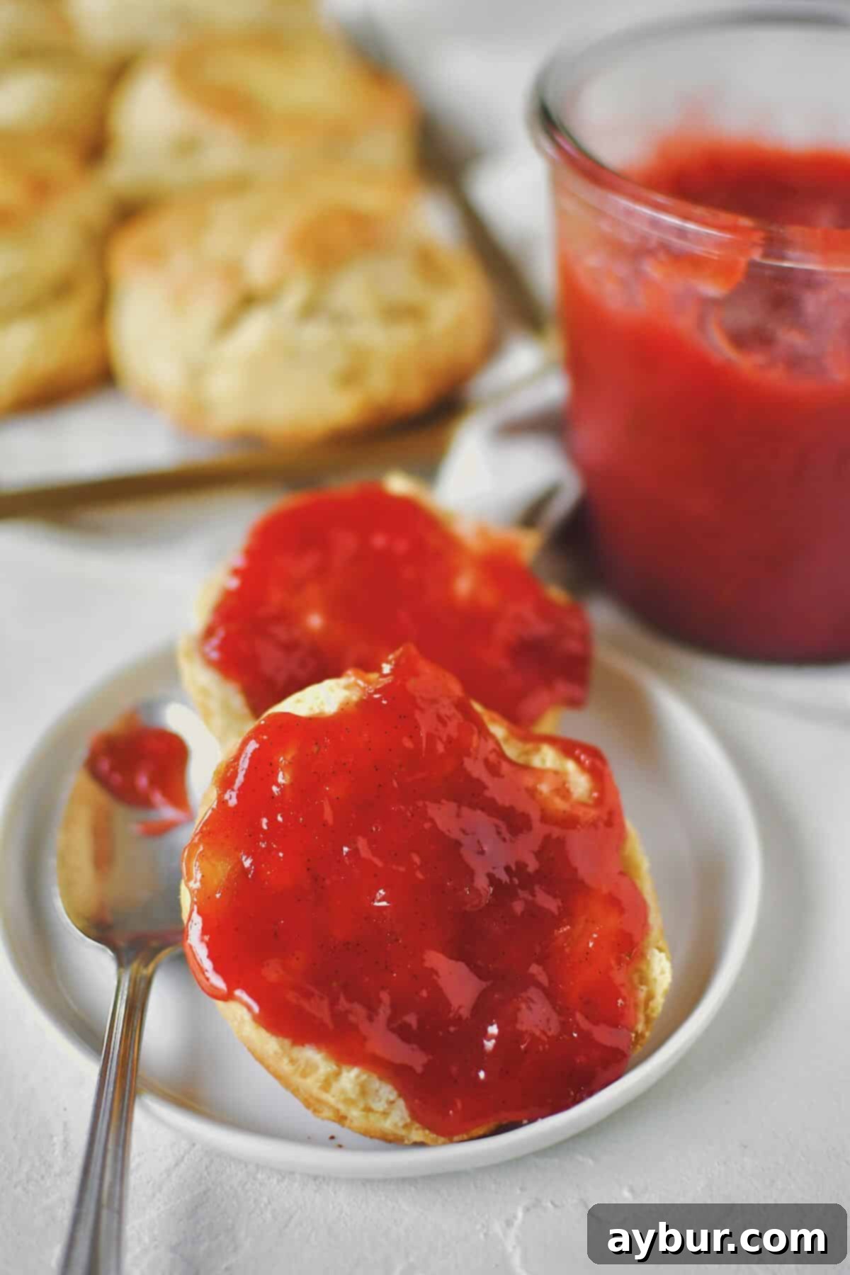 Sun-Kissed Strawberry Rhubarb Spread 12 A close-up of delicious Strawberry Rhubarb Jam spread generously on a freshly baked, flaky biscuit, with a small spoon resting beside it, inviting you to take a bite.