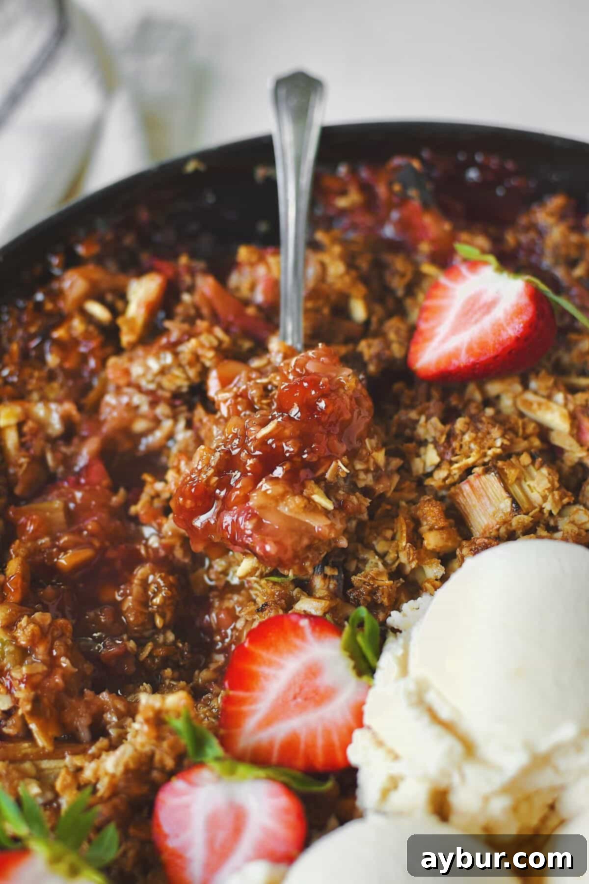 A close-up of a spoon scooping into a warm Strawberry Rhubarb Crisp, topped with melting ice cream and fresh strawberries, capturing a perfect first bite.