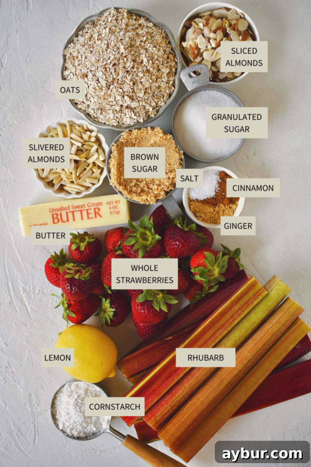 A flat lay of all the fresh ingredients needed to prepare a delicious Strawberry Rhubarb Crisp, including fresh strawberries, sliced rhubarb, lemon, sugar, cornstarch, oats, brown sugar, butter, and almonds.