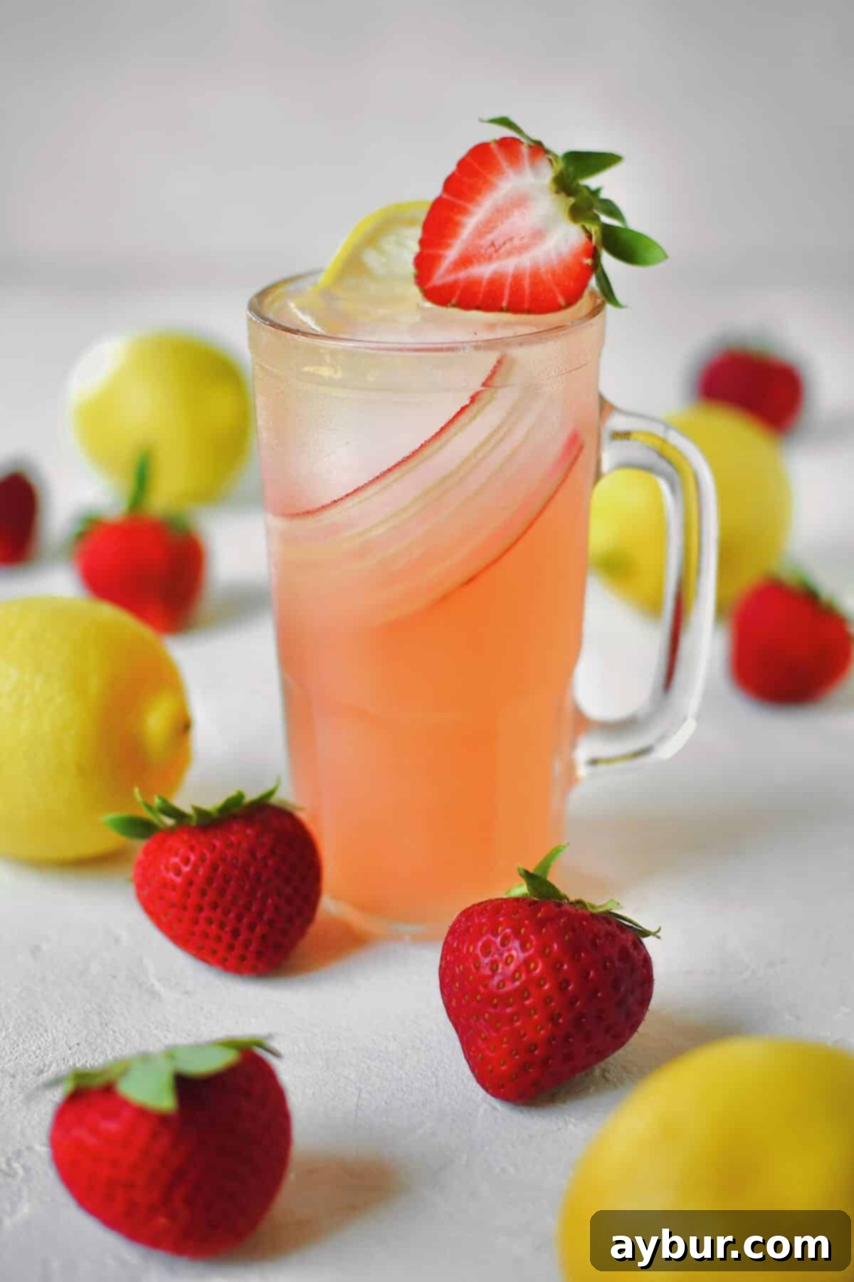 Sweet Strawberry Lemonade 6 A glass of strawberry lemonade on a table with fresh strawberries and lemons around it, garnished with a rhubarb curl.