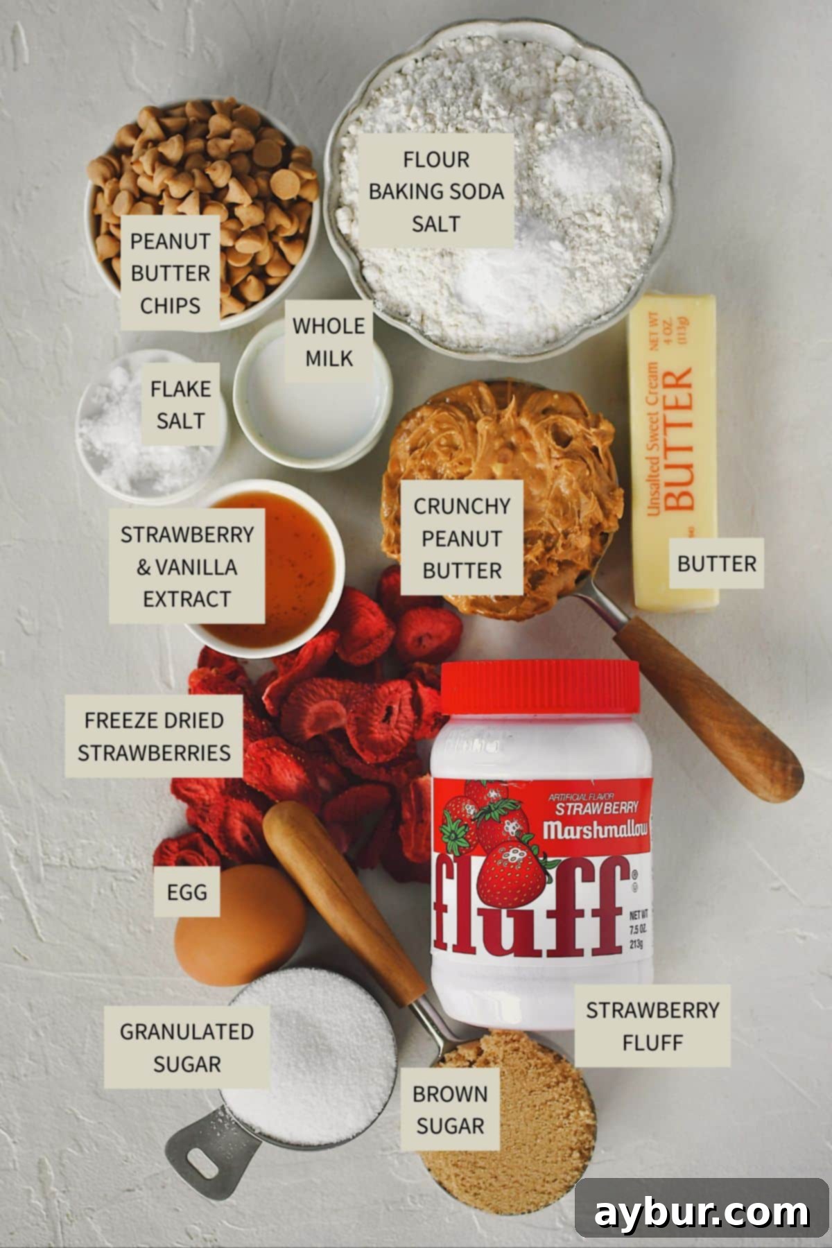 A collection of ingredients laid out for making Strawberry Fluffernutter Cookies, including peanut butter, butter, sugars, eggs, extracts, flour, baking soda, milk, freeze-dried strawberries, peanut butter chips, and strawberry marshmallow fluff.