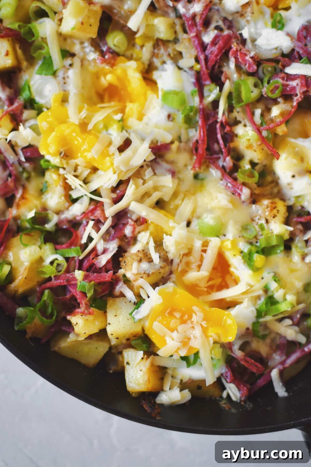 A close-up of the hearty corned beef hash, garnished with fresh green onions and melting cheese, highlighting the perfectly set egg whites and inviting runny yolks.