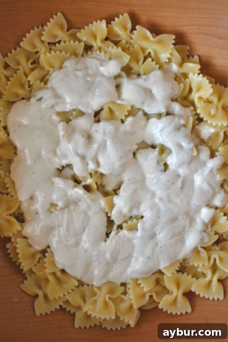 Cooked and cooled bow-tie pasta being tossed with creamy Caesar dressing in a large bowl, ensuring even coating.