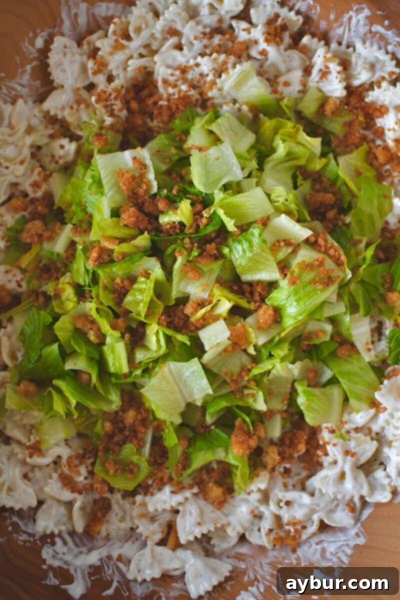 Adding crisp chopped romaine lettuce and toasted parmesan bread crumbs to the dressed pasta in a large mixing bowl for Caesar Pasta Salad.