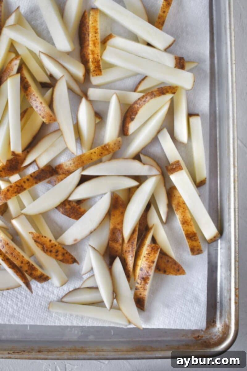 Freshly cut russet potato frites, spread out on a paper towel-lined baking sheet to dry completely after soaking, ensuring maximum crispness.