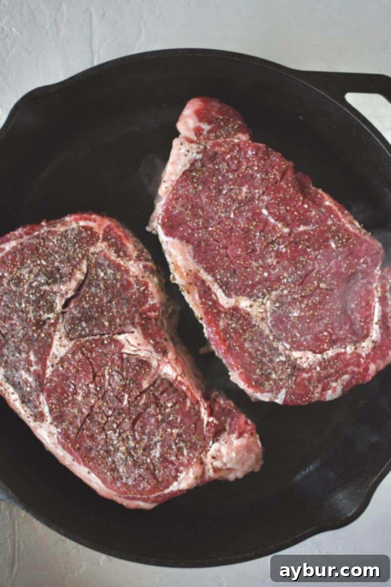 Thick-cut ribeye steaks, seasoned generously, being placed into a smoking hot cast iron pan to develop a rich, dark crust on the first side.