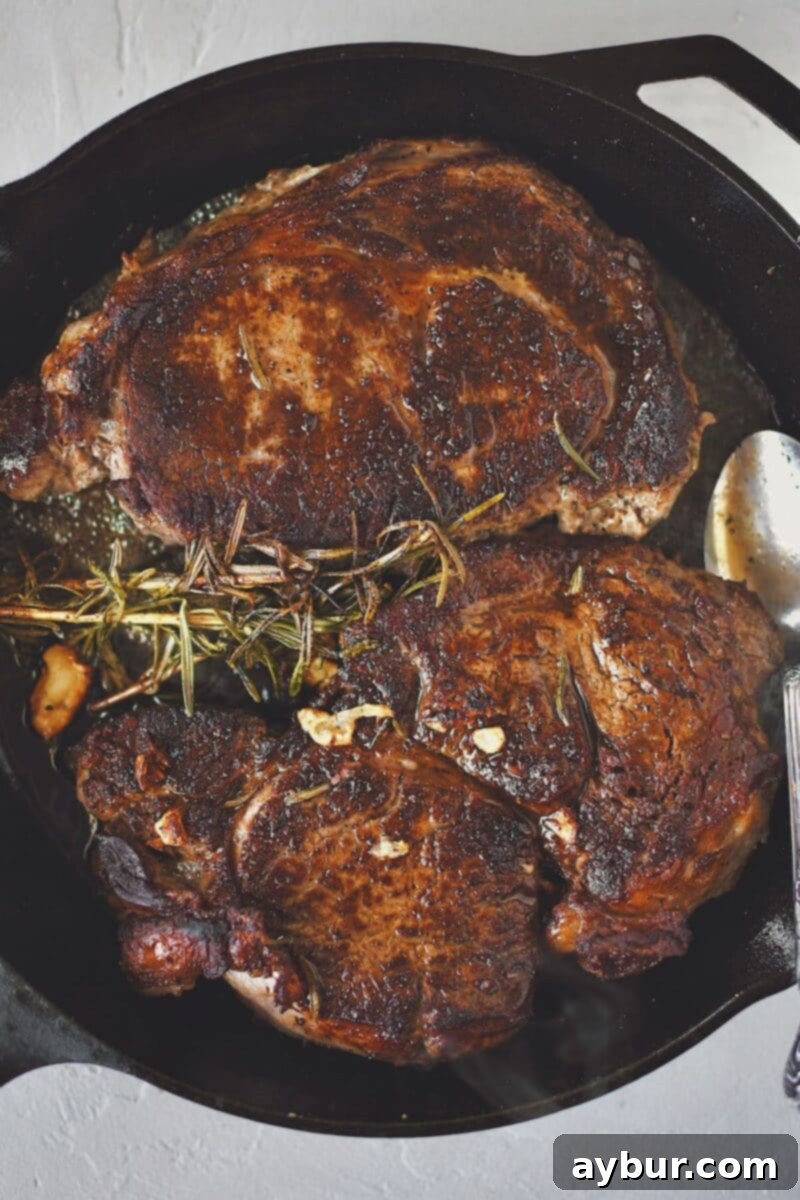 Beautifully seared ribeye steaks resting in the cast iron skillet, having been basted with butter, fragrant garlic, and fresh rosemary sprigs.