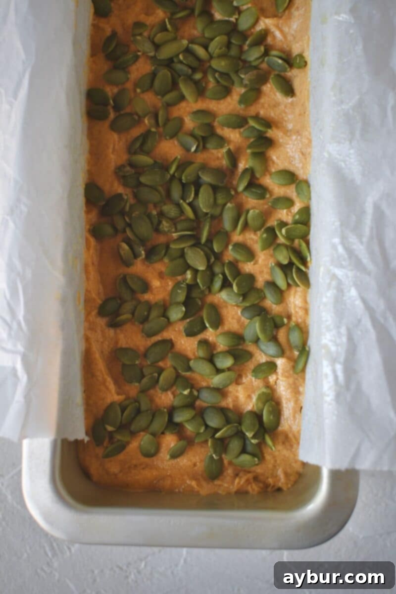 The pumpkin spice bread batter evenly spread in a loaf pan, topped with raw pepitas, ready for the oven.
