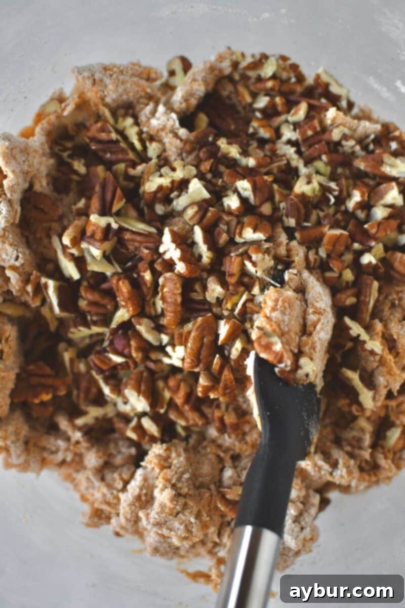 A visual of the toasted and chopped pecans being carefully folded into the pumpkin scone dough using a spatula, ensuring even distribution throughout the mixture.
