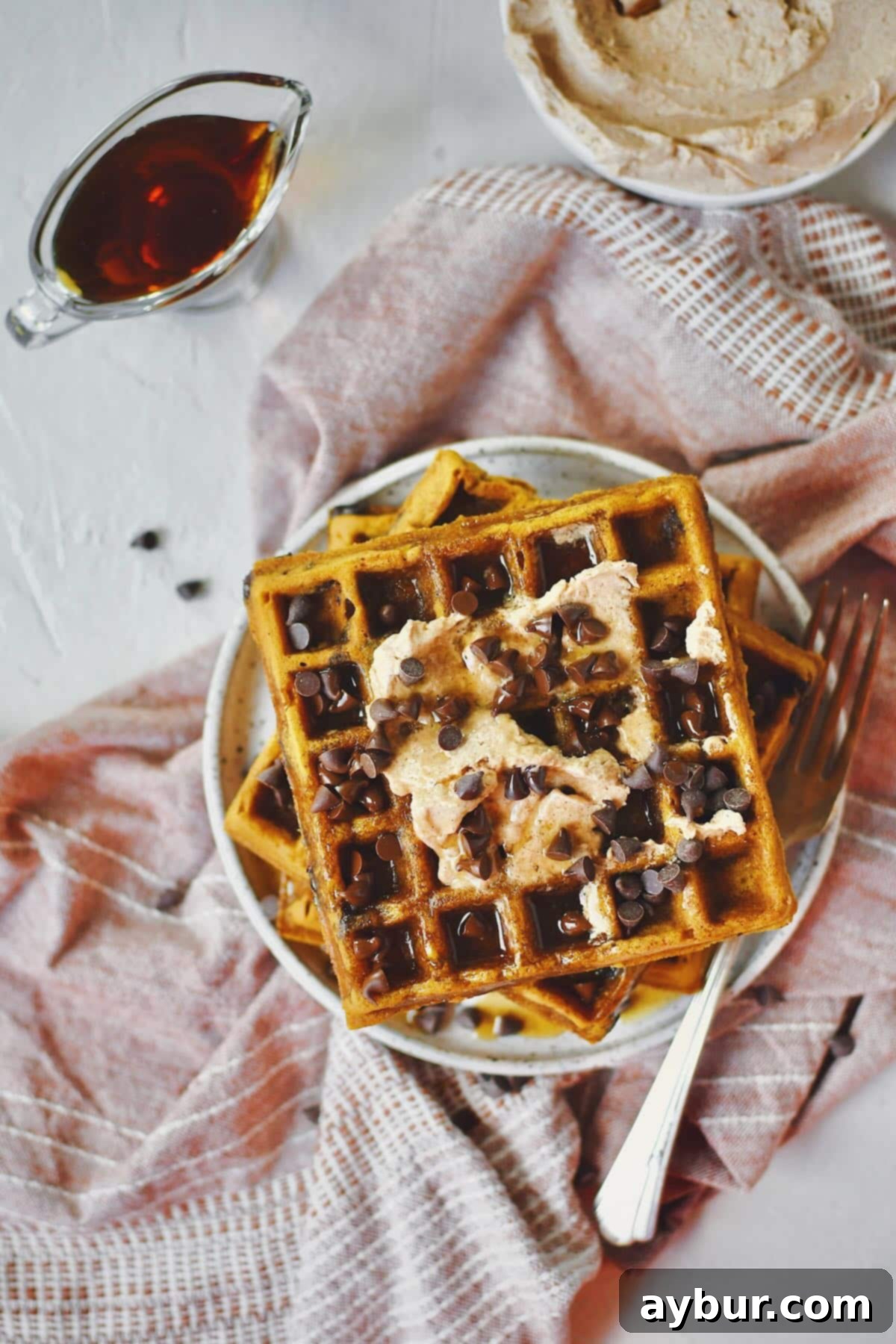 A stack of three perfectly cooked pumpkin waffles garnished with a small pat of cinnamon butter, a drizzle of maple syrup, and a generous sprinkle of chocolate chips on a white plate.