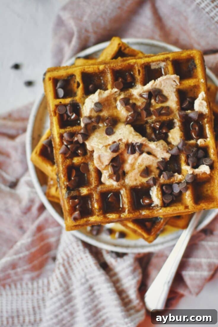 A stack of golden Pumpkin Waffles on a plate, generously topped with melted cinnamon butter, rich maple syrup, and a sprinkle of extra chocolate chips, ready to be enjoyed.