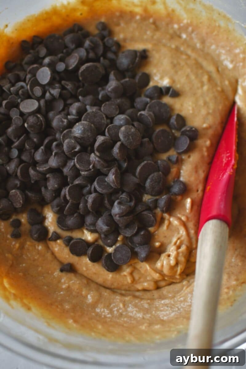 A close-up shot of semi-sweet chocolate chips being folded into the smooth pumpkin waffle batter with a spatula, distributing them evenly.